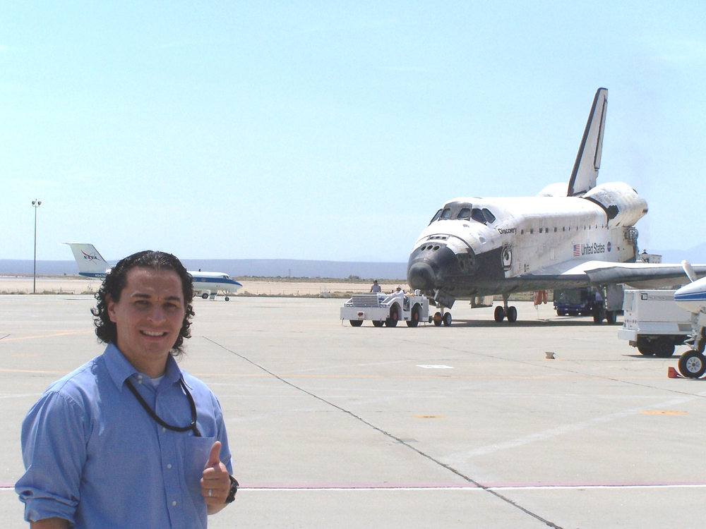 A person in a blue button down outside in daylight giving a thumbs up in front of a plane.